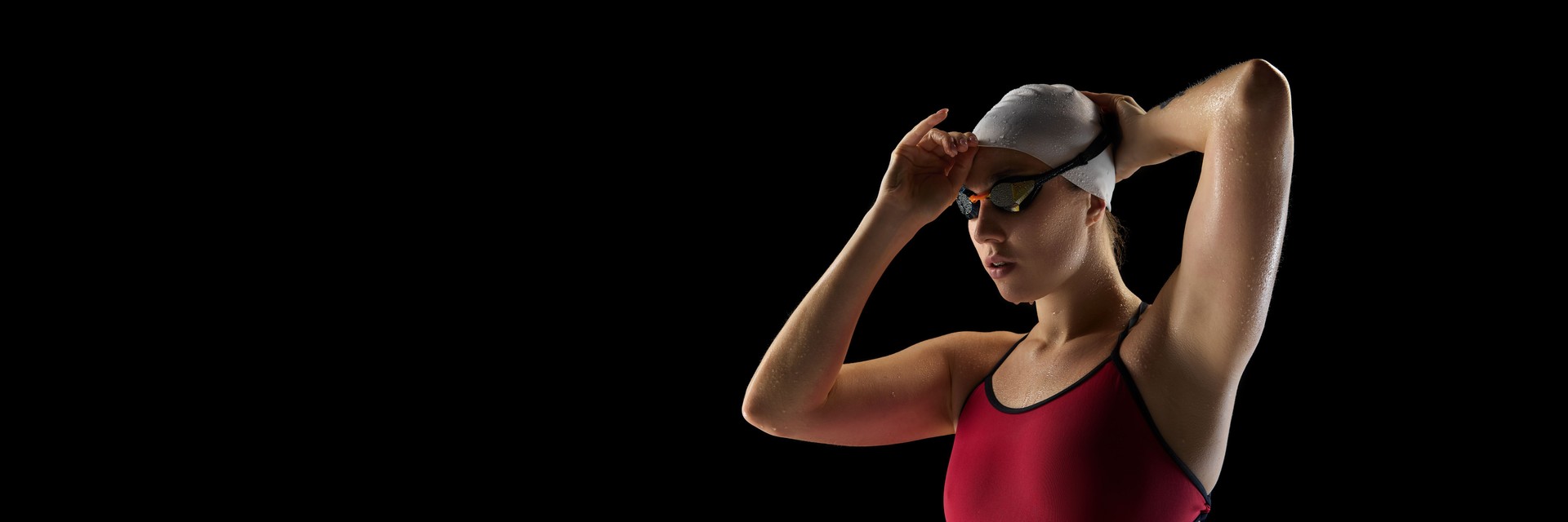 Banner. Athletic of young female swimmer wearing cap and goggles before swim race against black studio background. Negative space.