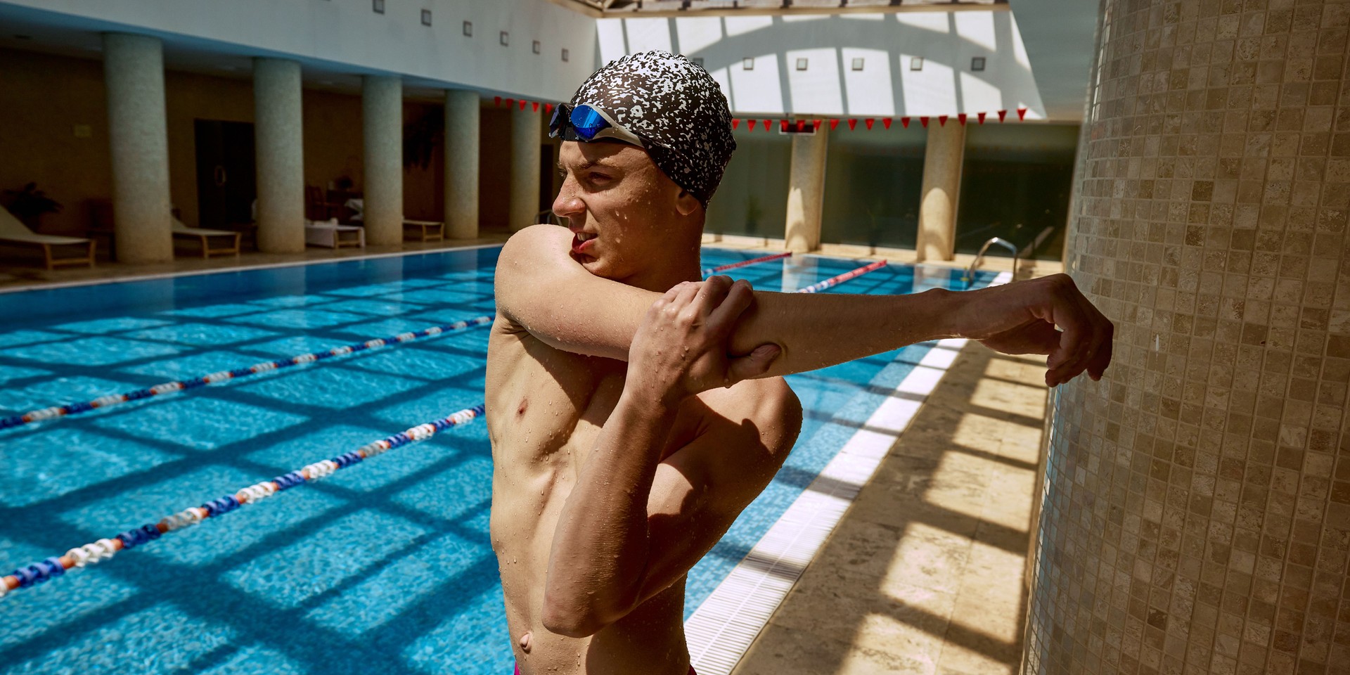 Young swimmer stretching arm beside indoor pool under natural light